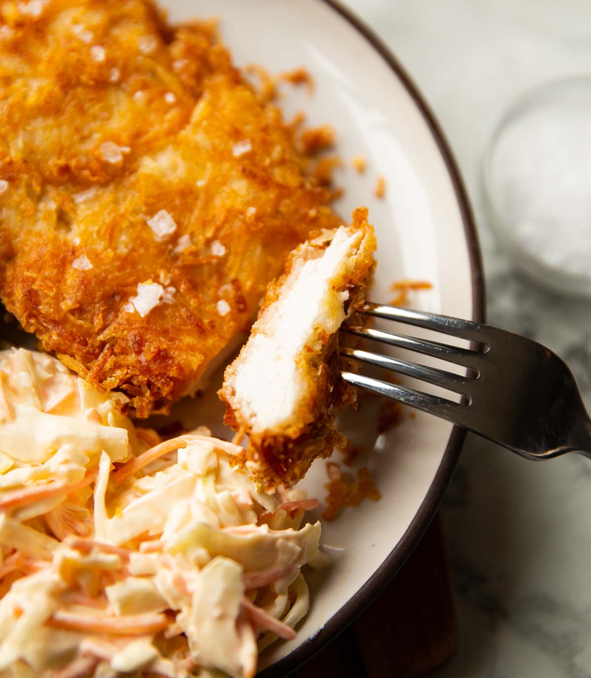 close up shot of silver fork cutting potato crusted chicken on small white plate with coleslaw